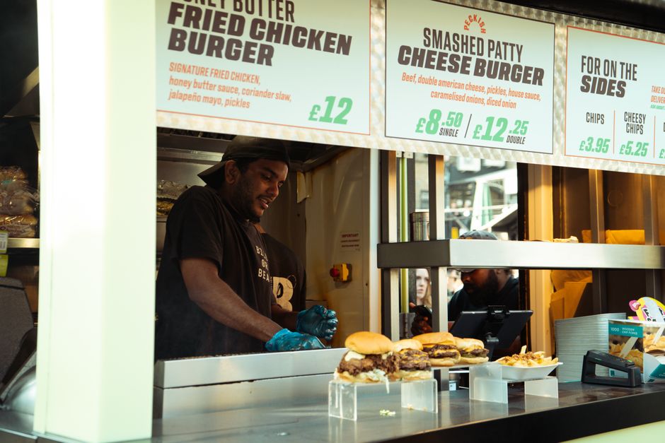 Inside the Kitchen area of Boxpark Shoreditch, a commercial space used for market trading, a young male staff member is preparing food behind a stainless steel counter, which is equipped with various 