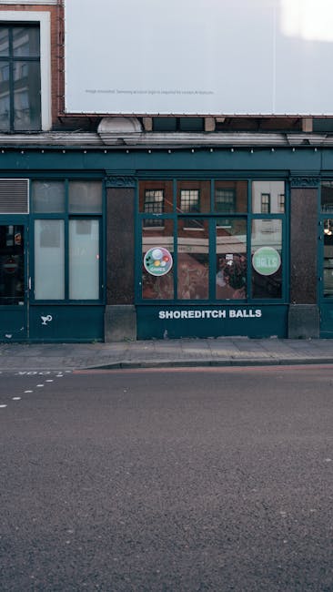 The exterior facade of Shoreditch Balls, a commercial building with a dark brick ground floor and large rectangular glass windows featuring circular signs that advertise sales and events. The windows